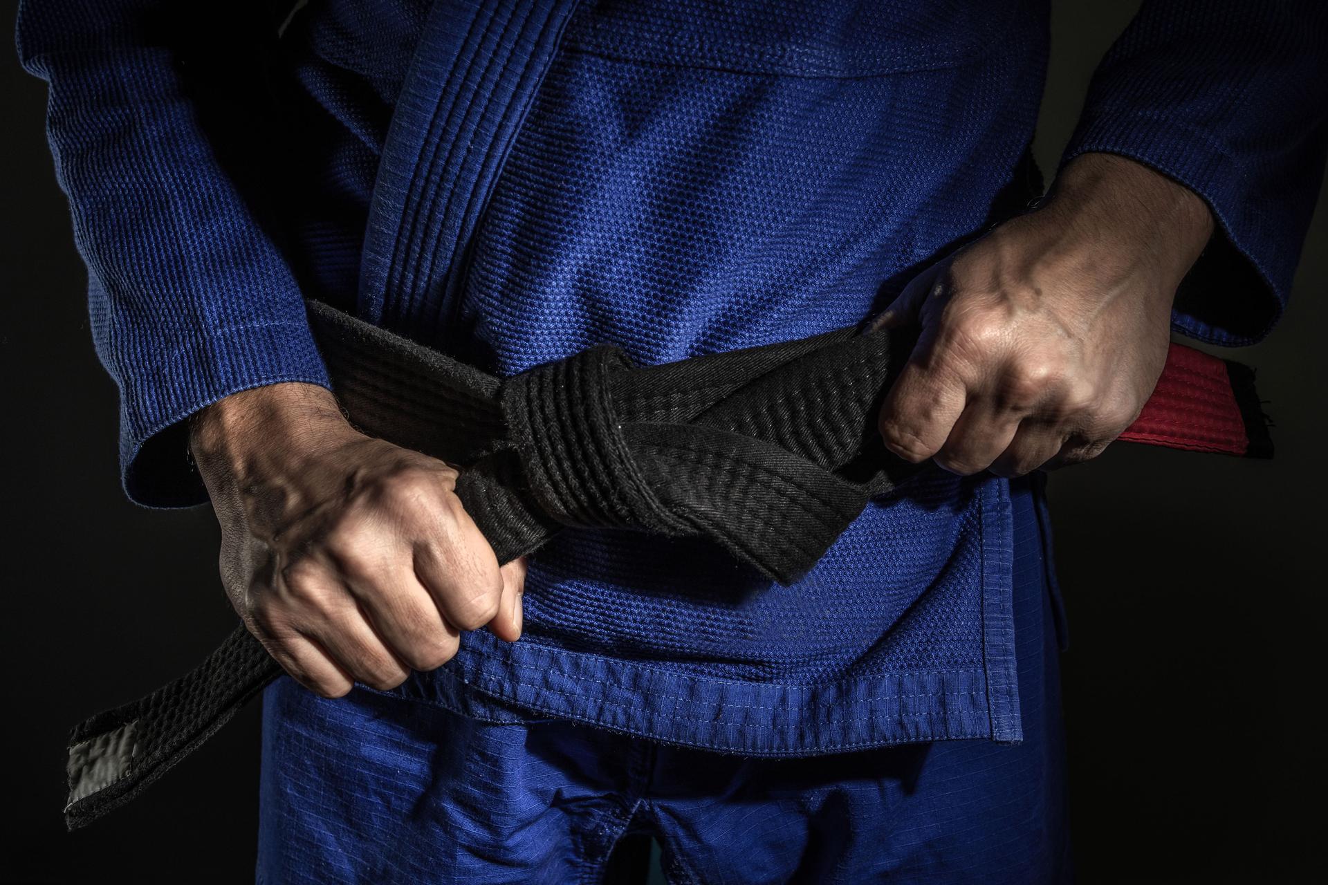 Close up on hand of unknown caucasian man holding brazilian jiu jitsu bjj black belt around his waist while wearing kimono gi in dark - martial arts mastery skill and confidence achievement concept