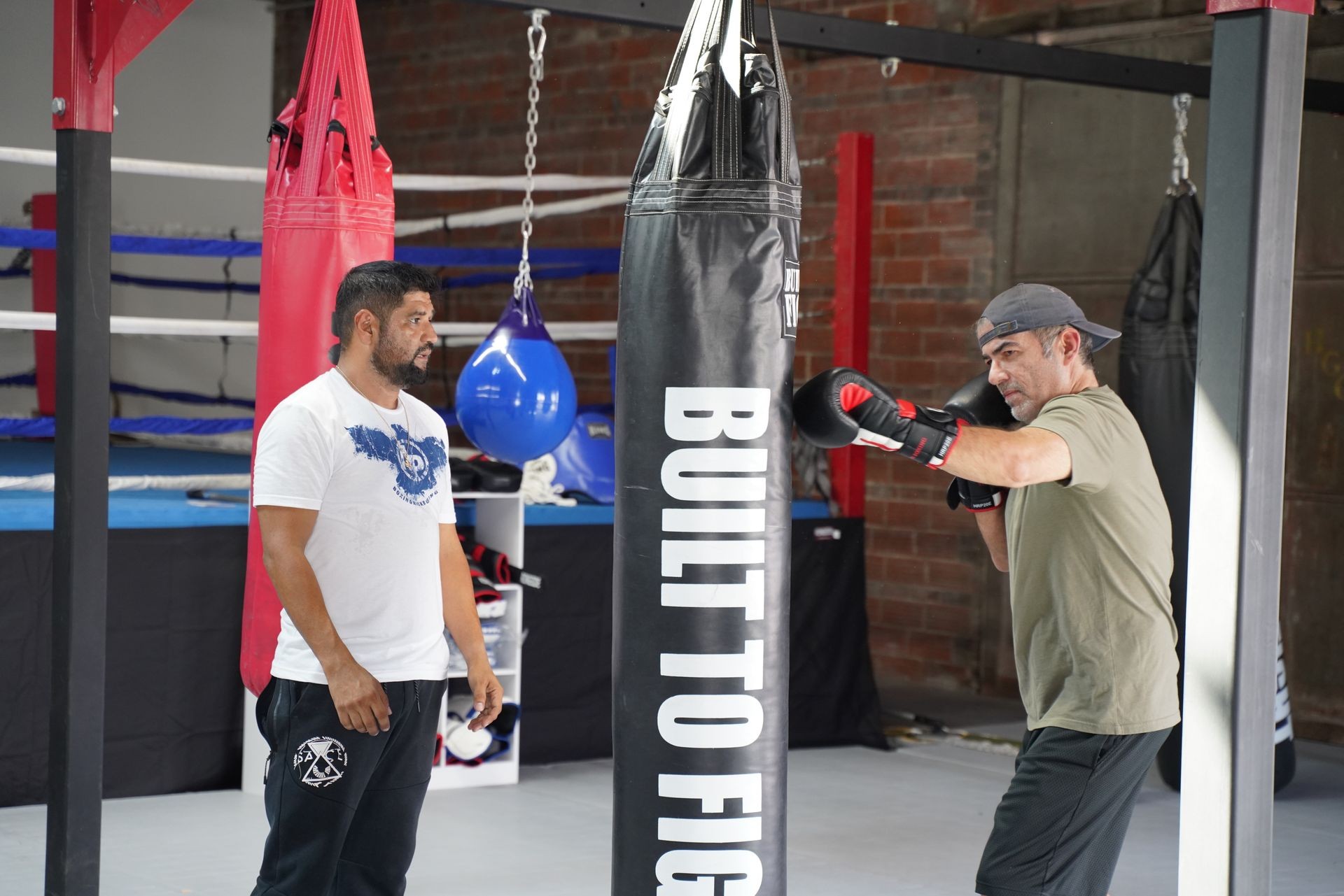 Man in boxing gloves training with a punching bag in a gym, with another man observing.